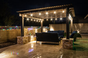 A resort-style backyard at night with a waterfall, pergola, and a firepit at night.
